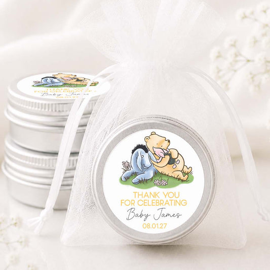 A round silver favor tin wrapped in a sheer white organza bag, with a custom round label featuring Winnie the Pooh and Eeyore. A stack of similar favor tins sits behind it on a soft white fabric background with delicate white floral accents.