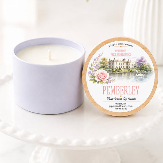 A soy candle in a matte container sits next to its round lid on a white pedestal. The lid features a printed label with a chateau illustration and the scent name 'Pemberley' in pink text, with white flowers and green leaves in the background.
