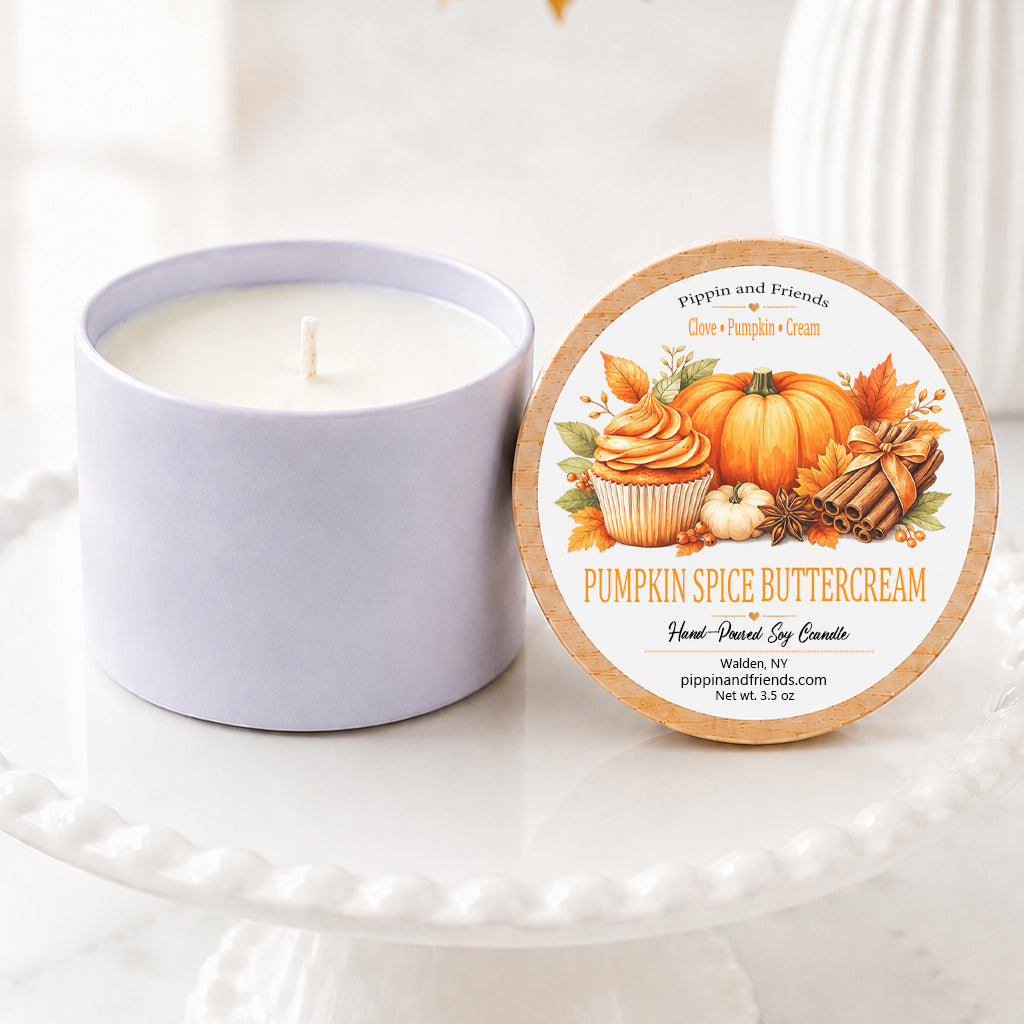 A soy candle in a white vessel sits on a white pedestal cake stand beside its round lid, which features a fall-themed illustration of pumpkin spice cupcakes and pumpkins. The background is softly blurred with autumn leaves and a white vase.
