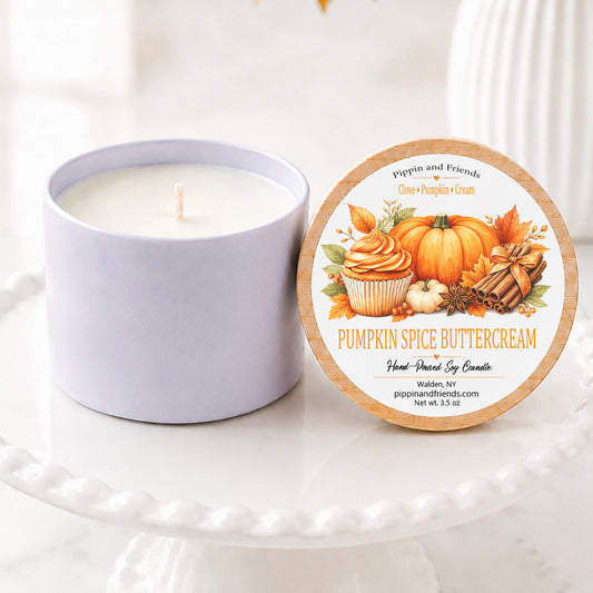 A soy candle in a white vessel sits on a white pedestal cake stand beside its round lid, which features a fall-themed illustration of pumpkin spice cupcakes and pumpkins. The background is softly blurred with autumn leaves and a white vase.