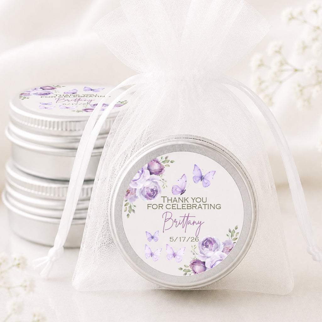 A small round silver tin with a purple floral and butterfly birthday favor label, tied in a sheer white organza drawstring bag, with two more favor tins stacked in the background on a soft white surface.