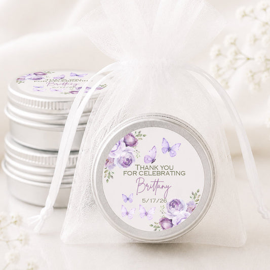 A small round silver tin with a purple floral and butterfly birthday favor label, tied in a sheer white organza drawstring bag, with two more favor tins stacked in the background on a soft white surface.