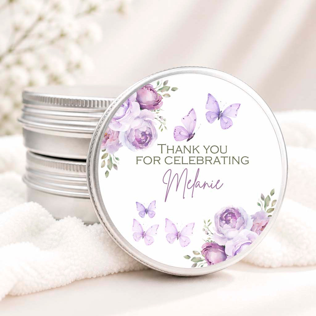 A stack of round silver metal tins with one tin open, showing a white lid printed with purple watercolor flowers, butterflies, and the text 'THANK YOU FOR CELEBRATING Melanie' in elegant script. The tins are styled on a soft white towel with blurred white flowers in the background.
