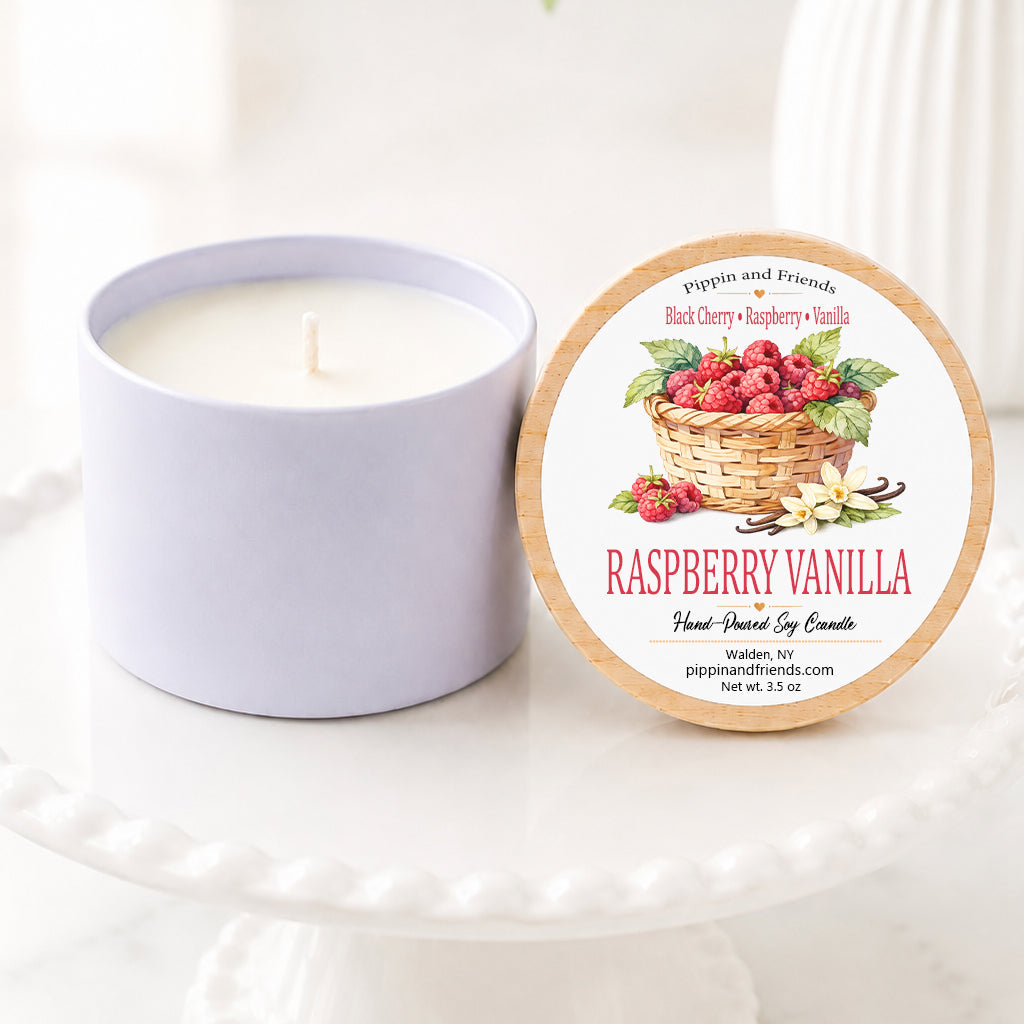 A  soy candle in a matte jar sits next to its round lid on a white pedestal. The lid features a label with a basket of raspberries and the text 'RASPBERRY VANILLA' and 'pippin and friends'.
