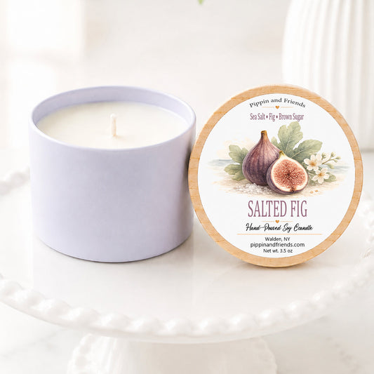 A soy candle in a matte container sits next to its round lid on a white pedestal. The lid features an illustration of a fig and the text 'SALTED FIG' and 'pippin and friends'.