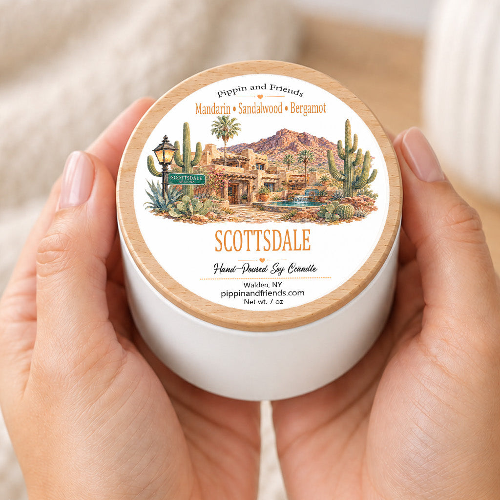 A person's hands holding a white jar candle with a round wooden lid. The lid features a detailed label for 'Scottsdale Hand-Shaped Soy Candle' by 'Pippin and Friends', depicting a desert landscape with cacti and a building.