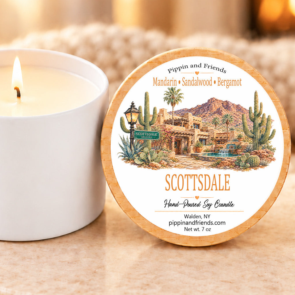 A round wooden-handled candle tin labeled 'SCOTTSDALE' with a desert landscape illustration, shown beside a lit white candle on a stone surface in a warm indoor setting.
