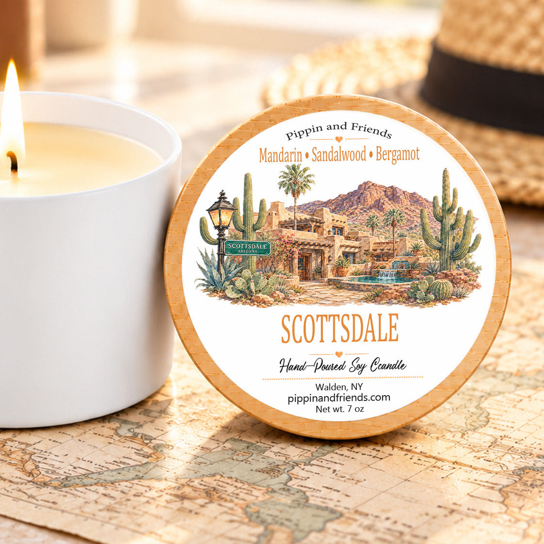A round wax candle tin labeled 'SCOTTSDALE' by Pippin and Friends sits on a vintage-style map next to a lit white candle. A woven hat is blurred in the background.