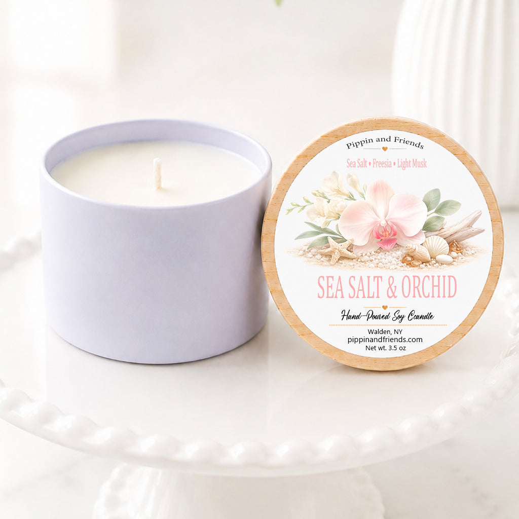 A sot candle in a matte cup sits next to its round lid on a white pedestal. The lid features a floral design with pink orchids and green leaves, labeled 'Sea Salt & Orchid' by 'pippin and friends'.