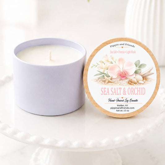 A sot candle in a matte cup sits next to its round lid on a white pedestal. The lid features a floral design with pink orchids and green leaves, labeled 'Sea Salt & Orchid' by 'pippin and friends'.
