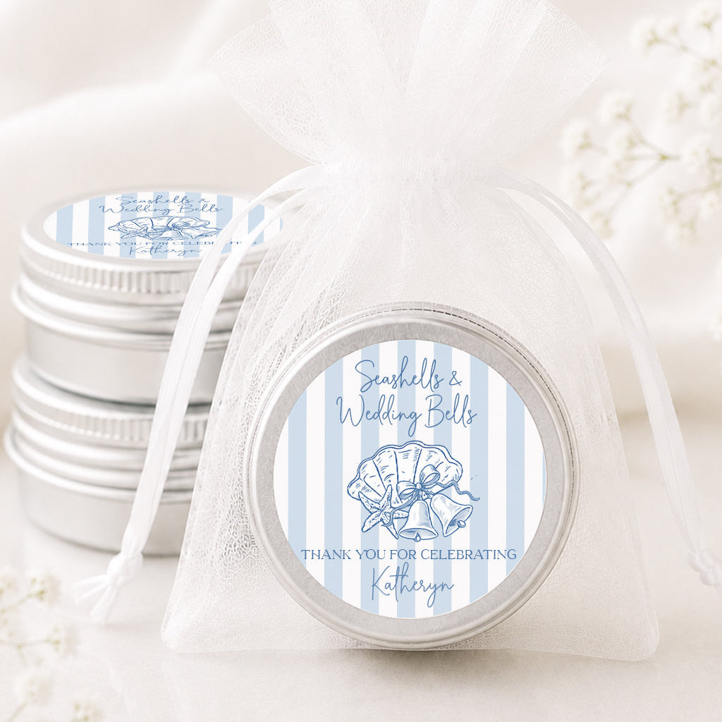 A small round silver favor tin tied with a sheer white organza bag, shown in front of a stack of similar favor tins. The front tin has a circular label with light blue and white vertical stripes, a seashell illustration, and the text 'Seashells Wedding Bells' and 'THANK YOU FOR CELEBRATING, Katherine'.