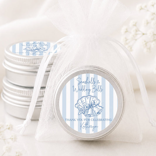 A small round silver favor tin tied with a sheer white organza bag, shown in front of a stack of similar favor tins. The front tin has a circular label with light blue and white vertical stripes, a seashell illustration, and the text 'Seashells Wedding Bells' and 'THANK YOU FOR CELEBRATING, Katherine'.