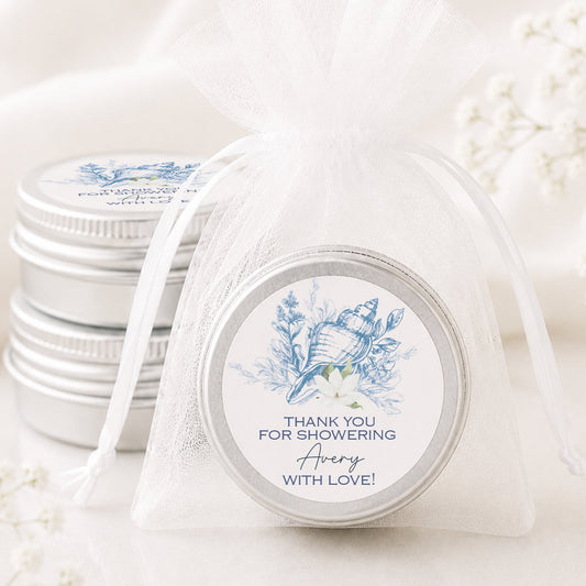A round silver tin wrapped in a sheer white organza drawstring bag, with a printed blue floral label that reads 'THANK YOU FOR SHOWERING Avery WITH LOVE!'. More matching silver tins are stacked in the background on a soft white surface with draped fabric and small white flowers.