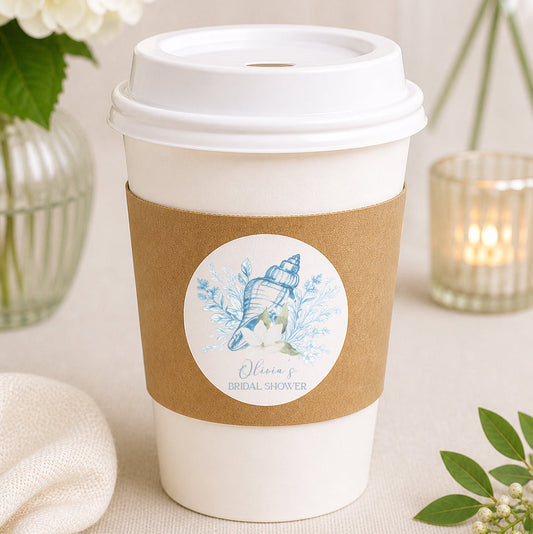 Elegant coastal bridal shower coffee cup favor featuring a personalized seashell sticker on a kraft sleeve with neutral wedding decor and soft floral accents.