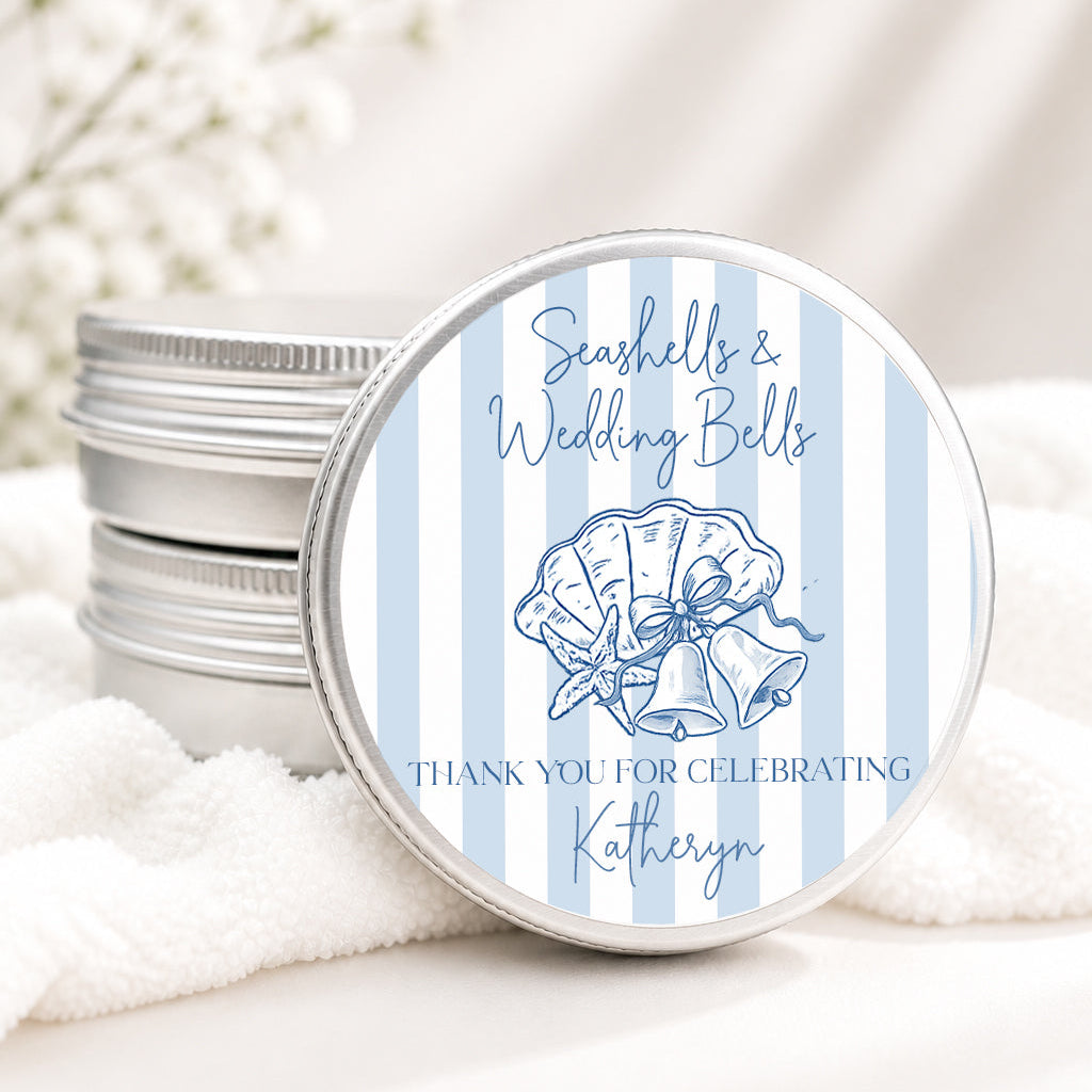 A round silver tin with a blue and white striped lid featuring a seashell illustration and the text 'Seashells & Wedding Bells' and 'Thank you for celebrating Kathryn,' shown on white fabric with a stack of similar tins in the background.