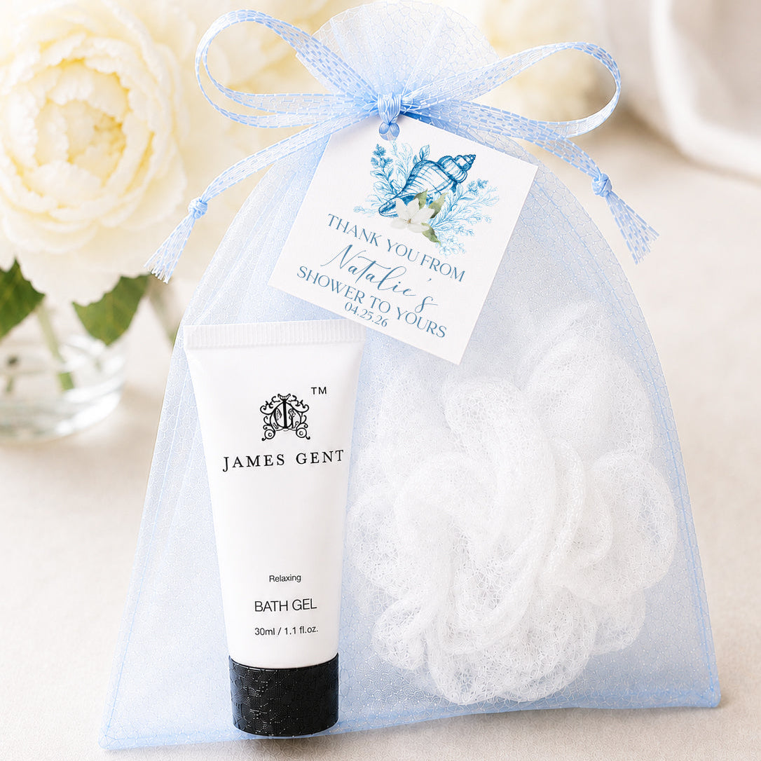 A gift bag containing a tube of James Gent bath gel and a white bath sponge with a personalized tag, placed on a table with white flowers in the background.