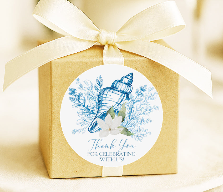 A brown gift box with a white ribbon and a round sticker attached to it. The sticker features a blue and white seashell and flowers, along with the text "Thank You for Celebrating With Us!"
