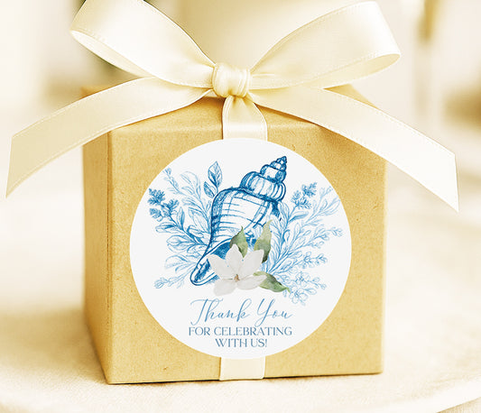 A brown gift box with a white ribbon and a round sticker attached to it. The sticker features a blue and white seashell and flowers, along with the text "Thank You for Celebrating With Us!"