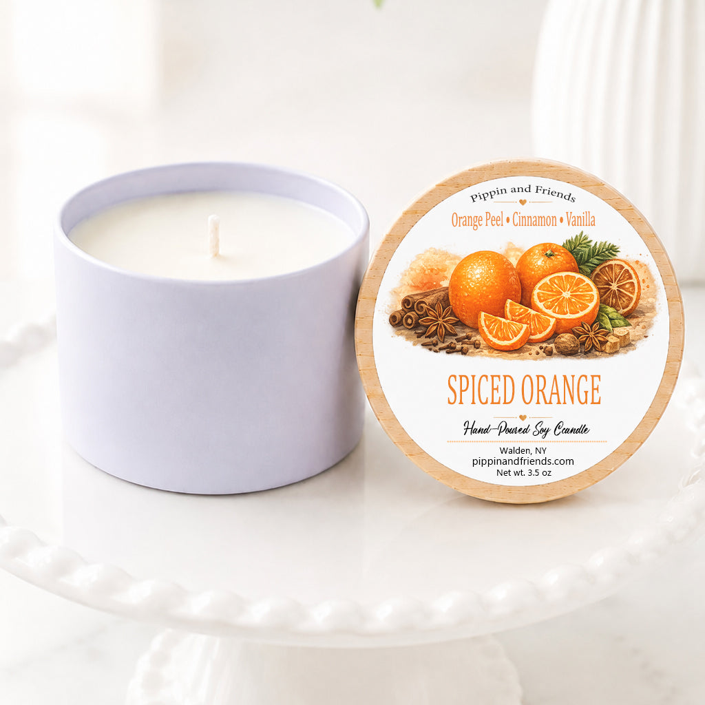 A soy candle in a matte jar sits next to its round lid on a white pedestal. The lid features a label for 'Spiced Orange' by Pippin and Friends, decorated with illustrations of oranges and spices.