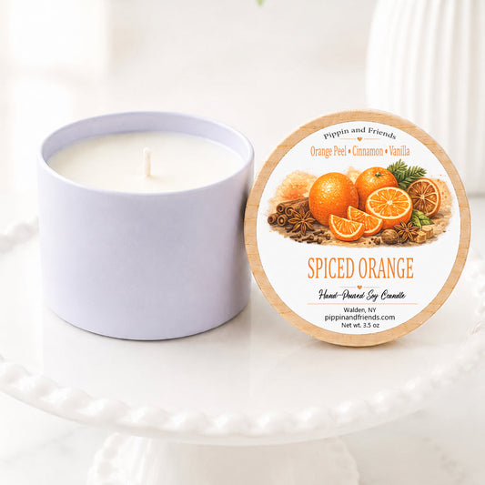 A soy candle in a matte jar sits next to its round lid on a white pedestal. The lid features a label for 'Spiced Orange' by Pippin and Friends, decorated with illustrations of oranges and spices.