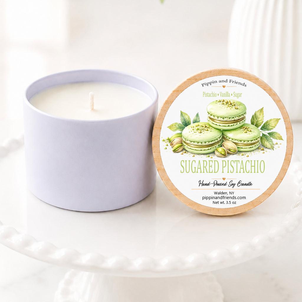 A soy candle in a matte cup sits next to its round lid on a white pedestal. The lid features a label for 'Sugared Pistachio' by 'pippin and friends' with illustrations of green pistachios and macarons.