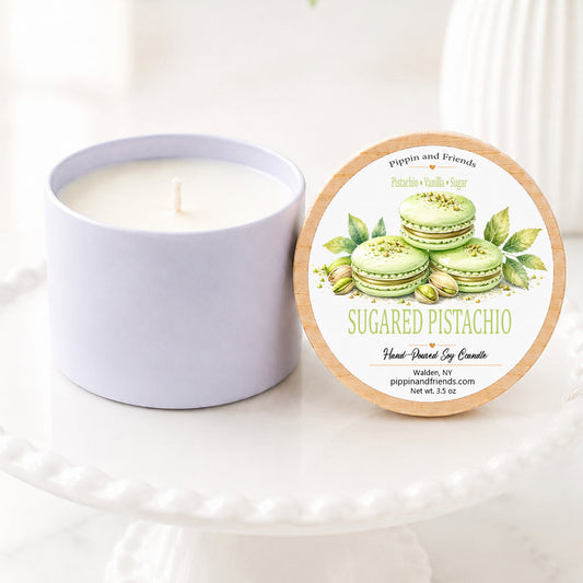 A soy candle in a matte cup sits next to its round lid on a white pedestal. The lid features a label for 'Sugared Pistachio' by 'pippin and friends' with illustrations of green pistachios and macarons.