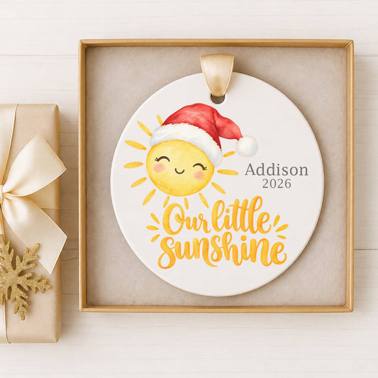 Personalized baby's first Christmas ceramic ornament featuring a cheerful watercolor sunshine wearing a red Santa hat, with “Our Little Sunshine” lettering, custom name Addison, and year 2026. Displayed with a satin ribbon in an elegant keepsake gift box.