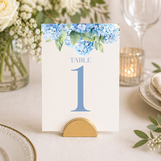 Blue hydrangea table number card displayed in gold stand on elegant wedding table setting with floral decor.