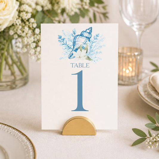 Elegant coastal seashell table number sign with blue watercolor shell artwork displayed in a gold holder on a neutral bridal shower reception table.