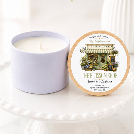 A soy candle in a matte cylinder container sits next to its round lid on a white pedestal. The lid features a natural wood rim and a detailed illustration of a flower shop named 'The Blossom Shop' by Pippin and Friends.