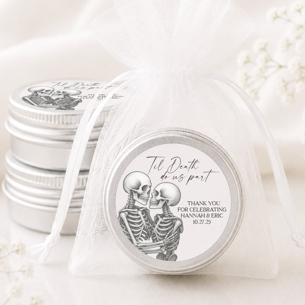 A small round silver favor tin sealed in a sheer white organza bag, with a printed skeleton illustration and the text 'The Death of us part' and 'THANK YOU FOR CELEBRATING JANNAMI & ERIC 8.2.