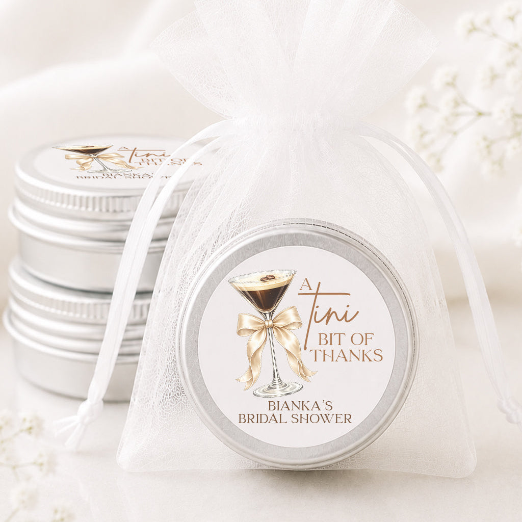 A small round silver tin wrapped in a sheer white organza bag, with a printed label that reads 'A tiny bit of thanks' and 'Bianca's Bachelorette.' Another stack of similar tins is visible in the background on a soft white surface.