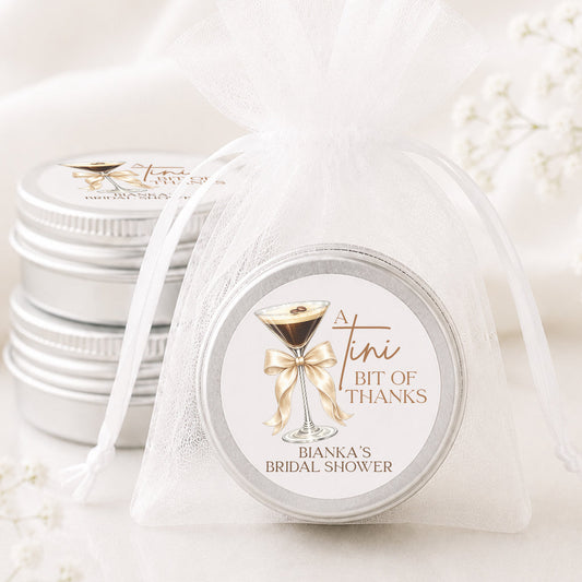 A small round silver tin wrapped in a sheer white organza bag, with a printed label that reads 'A tiny bit of thanks' and 'Bianca's Bachelorette.' Another stack of similar tins is visible in the background on a soft white surface.
