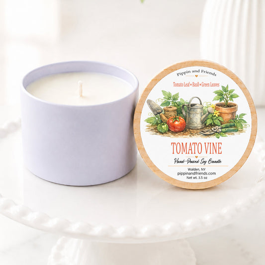 A soy candle in a matte container sits next to its round lid on a white pedestal. The lid features a natural wood border and an illustration of a tomato vine with gardening tools and herbs.