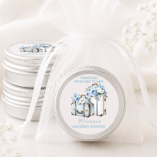 Personalized travel-themed wedding shower candle favor displayed in a white organza bag with a custom suitcase label, alongside stacked silver tin soy candles on a soft neutral background with delicate baby's breath flowers.