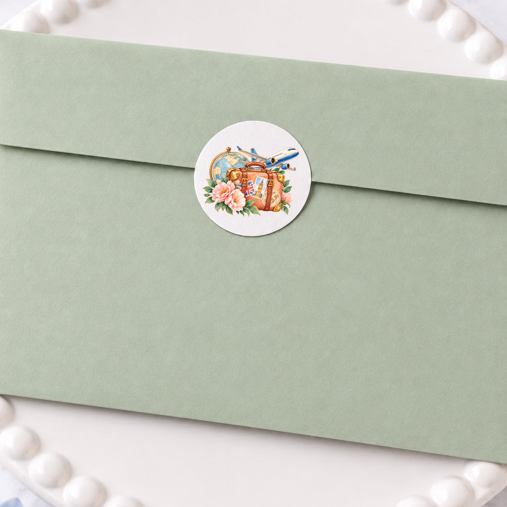 A sage green envelope with a round white sticker seal featuring a vintage illustration of a globe, flowers, and a suitcase, resting on a white beaded plate.