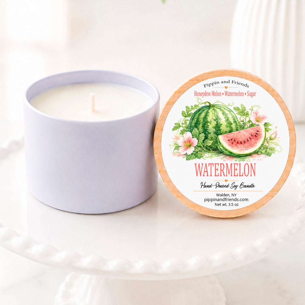 A soy candle in a matte cup sits next to its round lid on a white pedestal. The lid features a watermelon-themed illustration with the text 'Watermelon' and 'pippin and friends'.