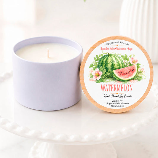 A soy candle in a matte cup sits next to its round lid on a white pedestal. The lid features a watermelon-themed illustration with the text 'Watermelon' and 'pippin and friends'.