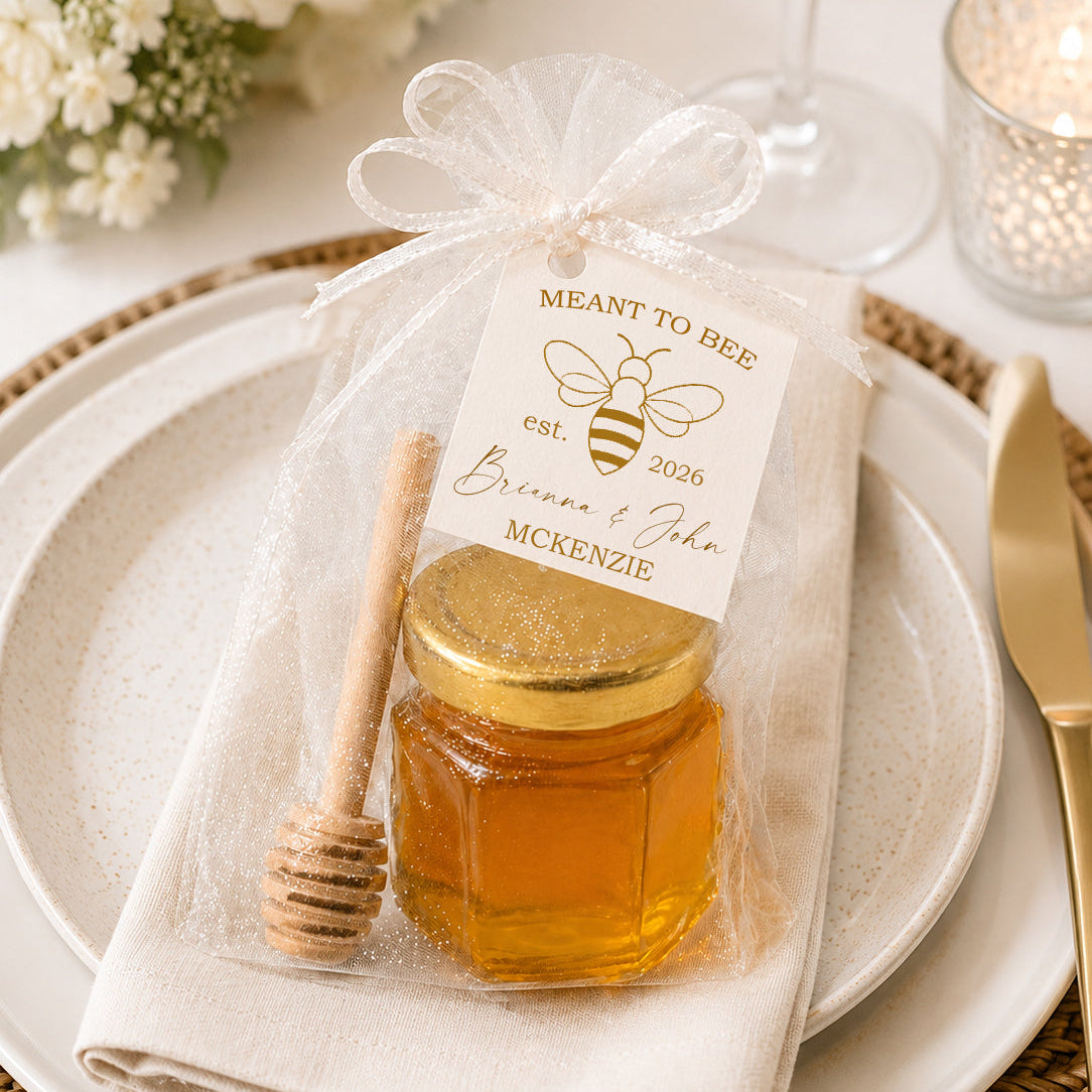 Honey jar wedding favor kit displayed on an elegant wedding place setting with a gold-lidded glass jar, wooden honey dipper, sheer white tulle bag, and personalized Meant to Bee wedding favor tag.