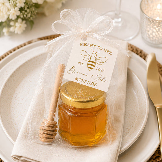 Honey jar wedding favor kit displayed on an elegant wedding place setting with a gold-lidded glass jar, wooden honey dipper, sheer white tulle bag, and personalized Meant to Bee wedding favor tag.