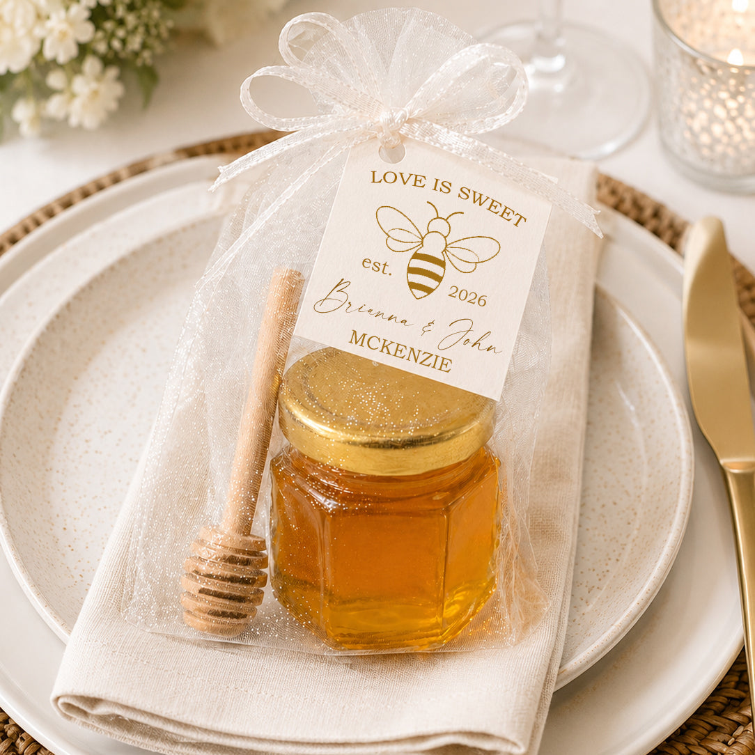 Love Is Sweet honey jar wedding favor kit displayed on an elegant wedding place setting with a mini glass jar filled with honey, gold lid, wooden honey dipper, sheer white tulle bag, and personalized bee-themed wedding tag.