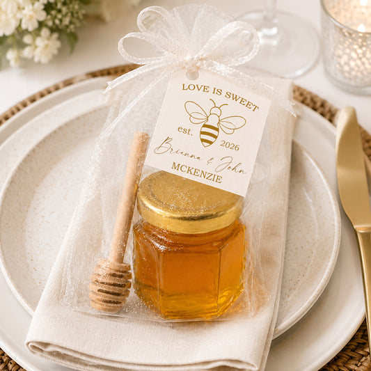 Love Is Sweet honey jar wedding favor kit displayed on an elegant wedding place setting with a mini glass jar filled with honey, gold lid, wooden honey dipper, sheer white tulle bag, and personalized bee-themed wedding tag.