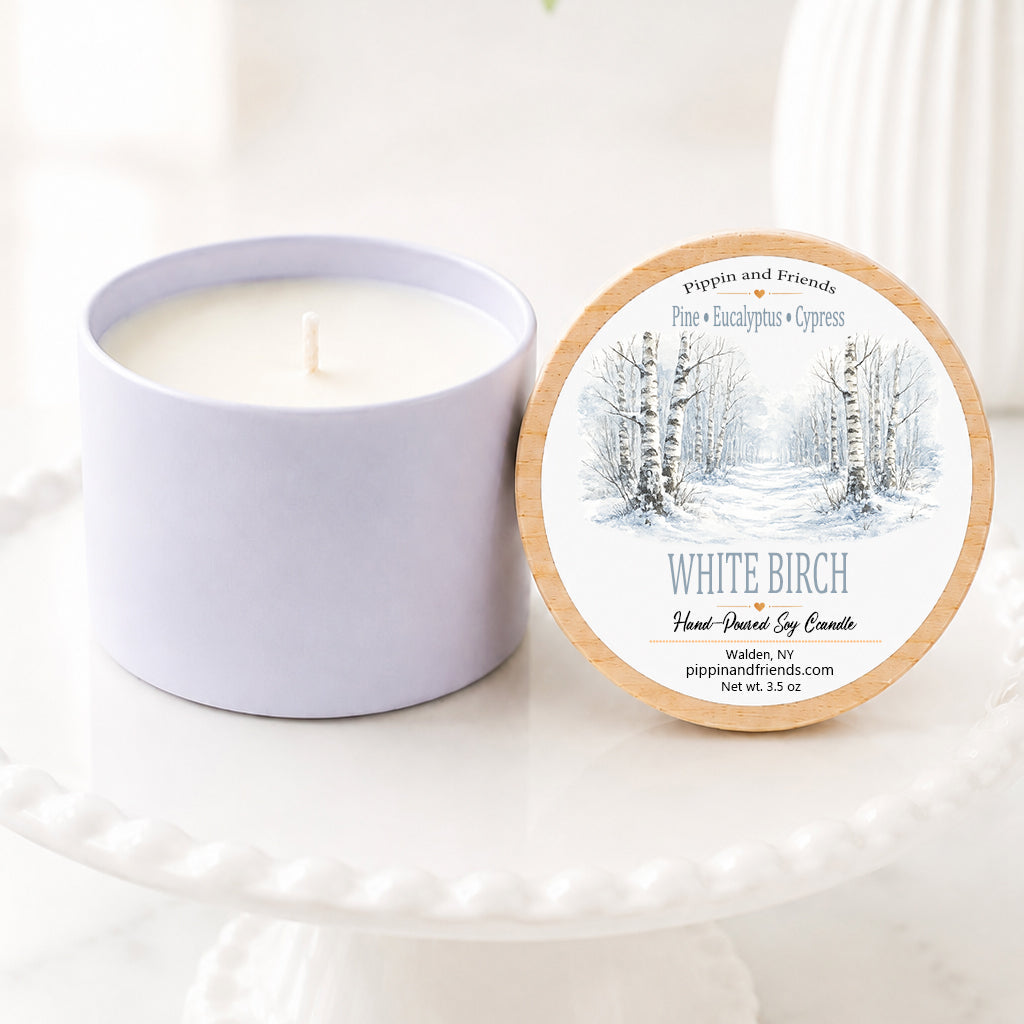 A soy candle in a matte container sits next to its round lid on a white pedestal. The lid features a winter forest illustration and the scent name 'White Birch'.