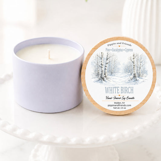A soy candle in a matte container sits next to its round lid on a white pedestal. The lid features a winter forest illustration and the scent name 'White Birch'.