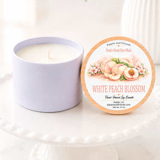 A soy candle jar next to its round lid, which features a peach and floral illustration and the text 'White Peach Blossom' by 'pippin and friends', displayed on a white pedestal stand.