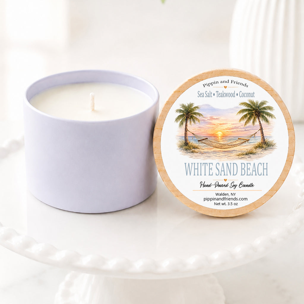 A soy candle in a matching cup sits beside its round lid on a white pedestal. The lid features a tropical beach illustration with palm trees and the text 'White Sand Beach' and 'Pipkin and Friends'.