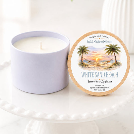 A soy candle in a matching cup sits beside its round lid on a white pedestal. The lid features a tropical beach illustration with palm trees and the text 'White Sand Beach' and 'Pipkin and Friends'.